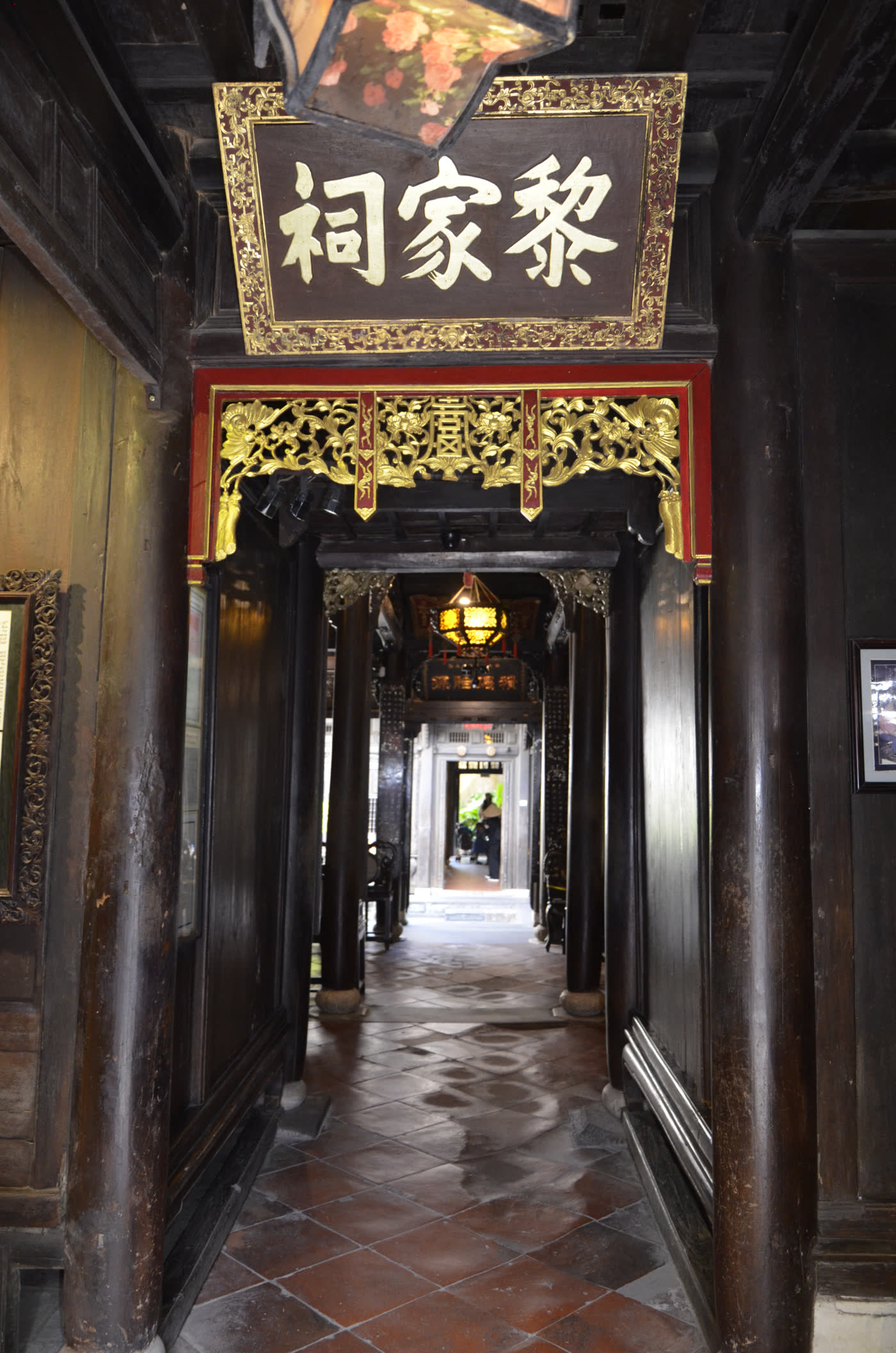 A board reading Lê Gia Từ in gilt Chinese characters hangs above a doorway inside Tấn Ký house, seen through several receding rows of dark timber columns toward a skywell at the back.