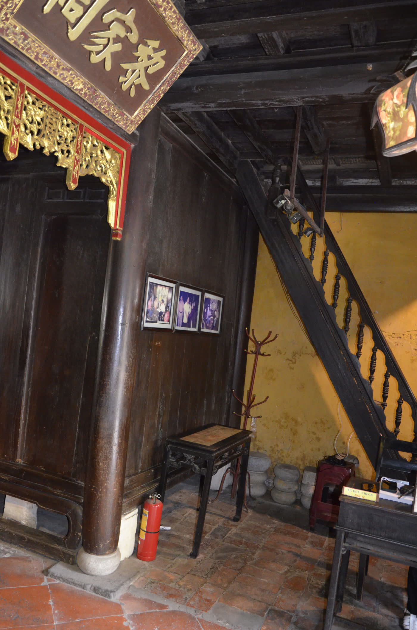 A wooden staircase inside Tấn Ký house rising beside round timber columns set on stone bases, tiled floor below, a red-lacquered gilt shrine board visible at upper left.