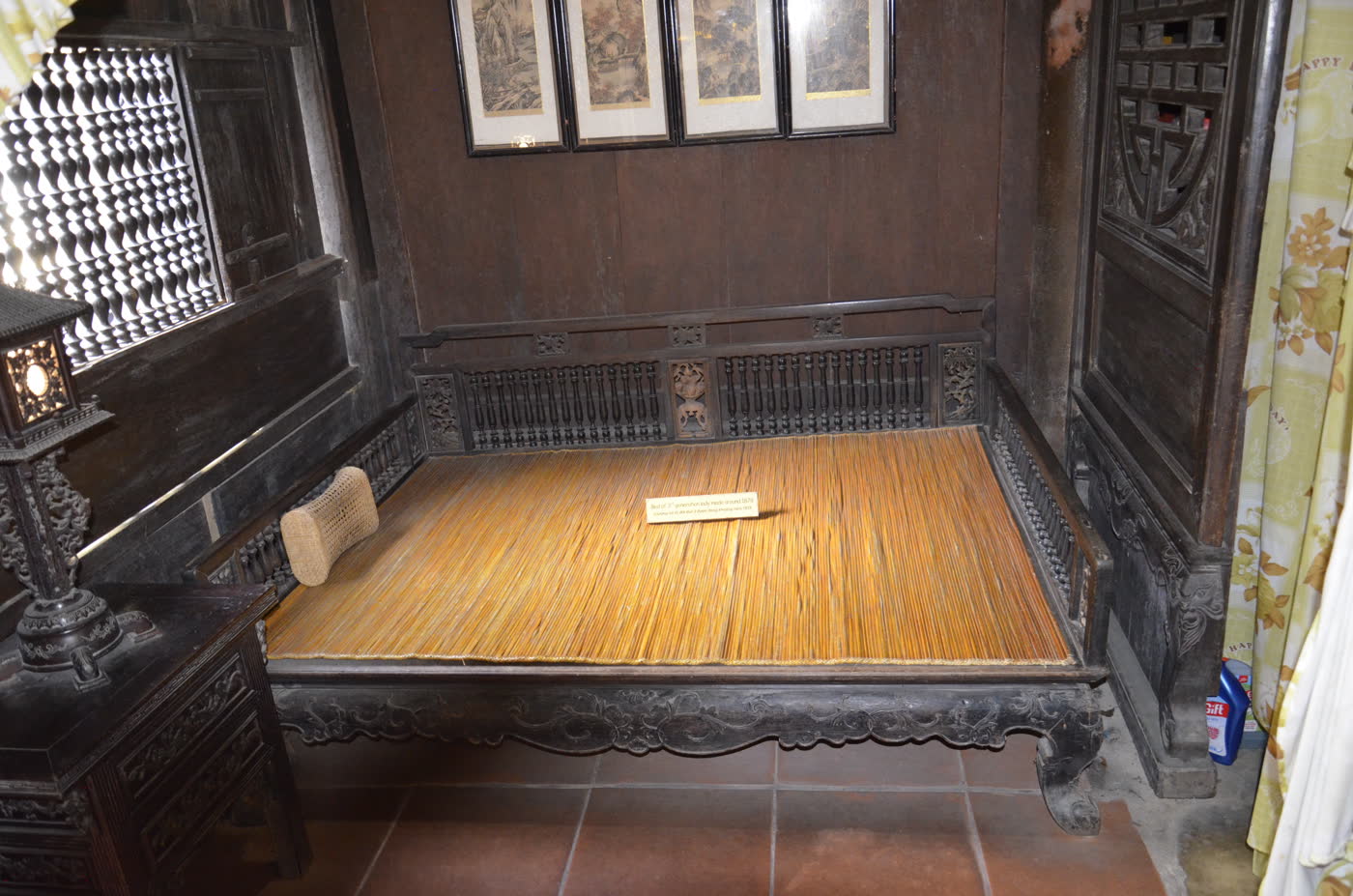 A carved dark wood daybed spread with a bamboo-slat mat and a cylindrical headrest, framed by a lattice window of coin-shaped openings on one side and a carved wooden panel on the other.
