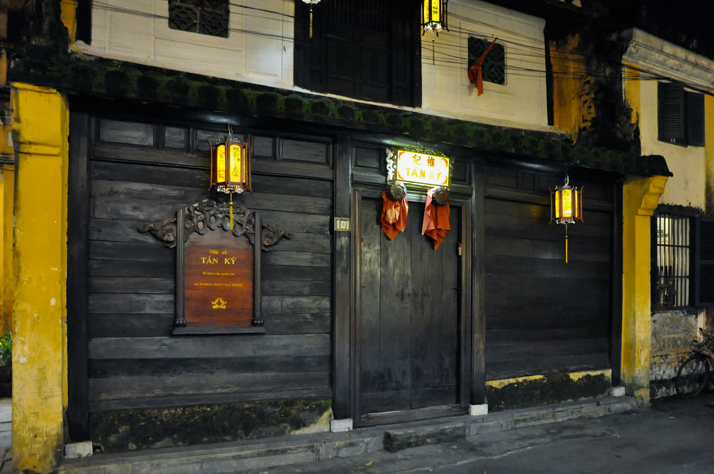 The closed wooden shopfront of Tấn Ký house at night, dark timber shutters and doors flanked by ochre-yellow walls, red lanterns lit on either side, a plaque reading TẤN KÝ above the door and a national-heritage-house sign mounted at lower left, the number 101 above the entrance.