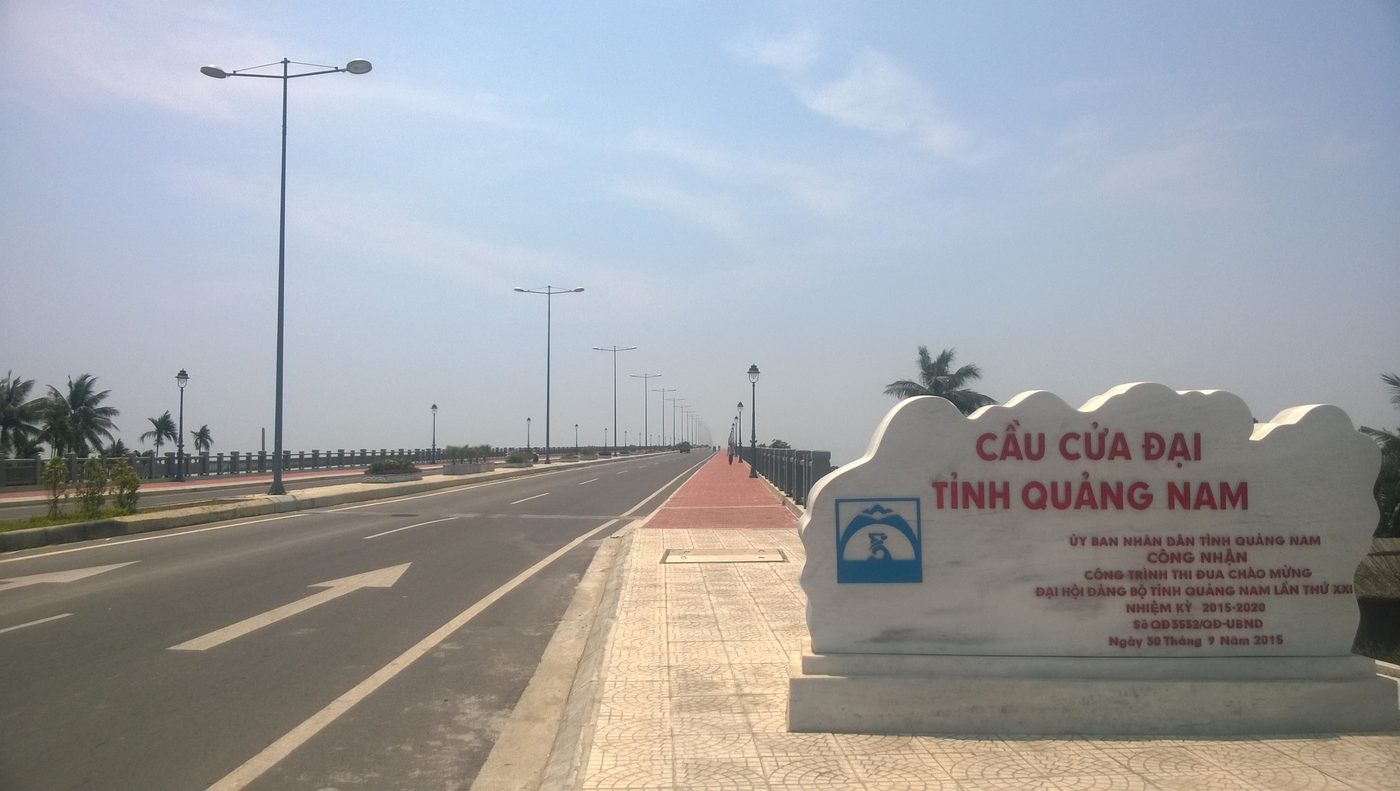 The Cửa Đại bridge across the lower Thu Bồn, a long concrete deck over a wide reach of water with low sandy banks on either side.