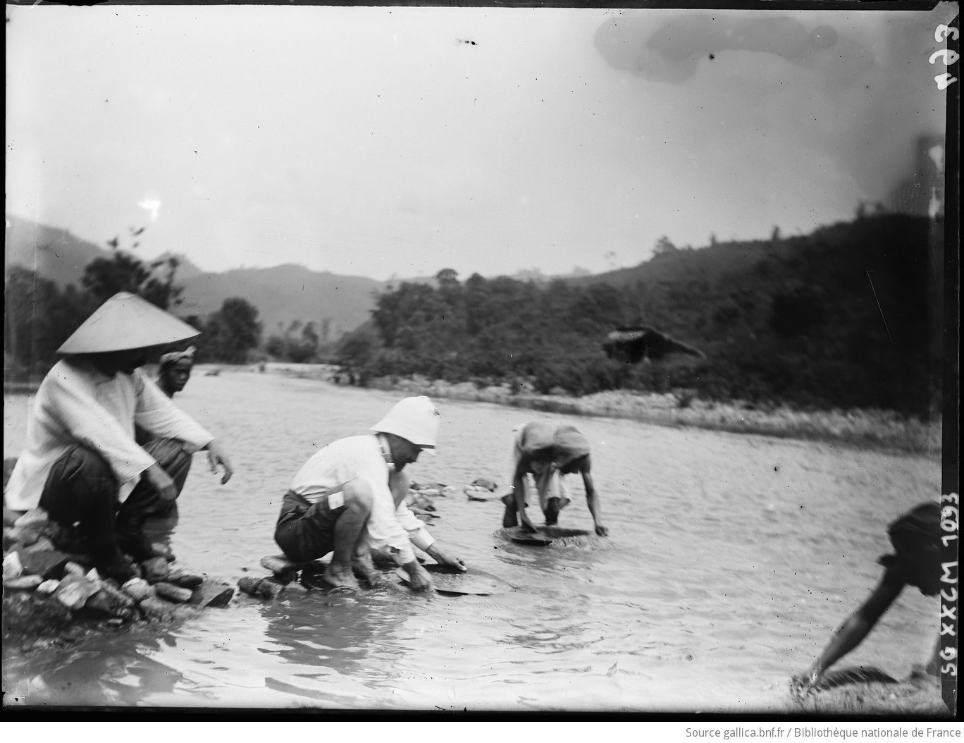 A black-and-white photograph from the colonial period: several men in conical hats and a Frenchman in a pith helmet wading in a mountain river, bent over round pans, working the gravel.