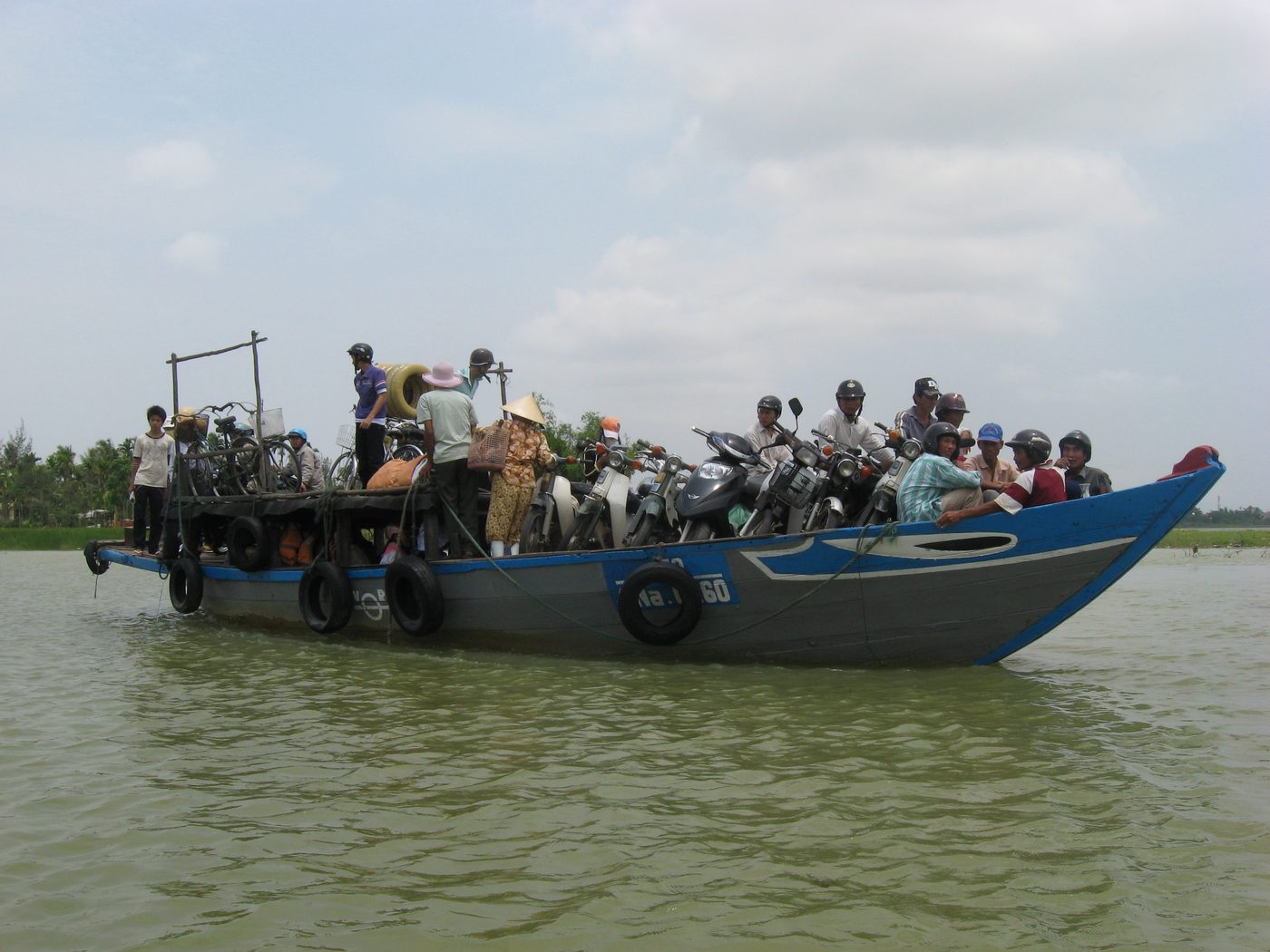 A ferry carrying people and motorbikes from Kim Bồng village across the Thu Bồn to Hội An, passengers sitting along the gunwale, the broad river behind.