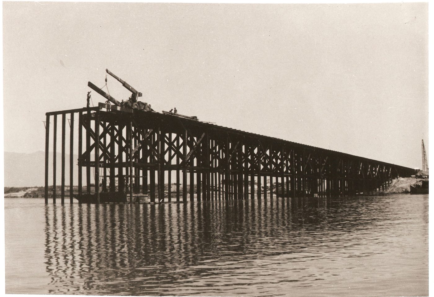 A timber trestle bridge under construction across the Thu Bồn, a crane on the deck lowering a beam, a few men standing on it.