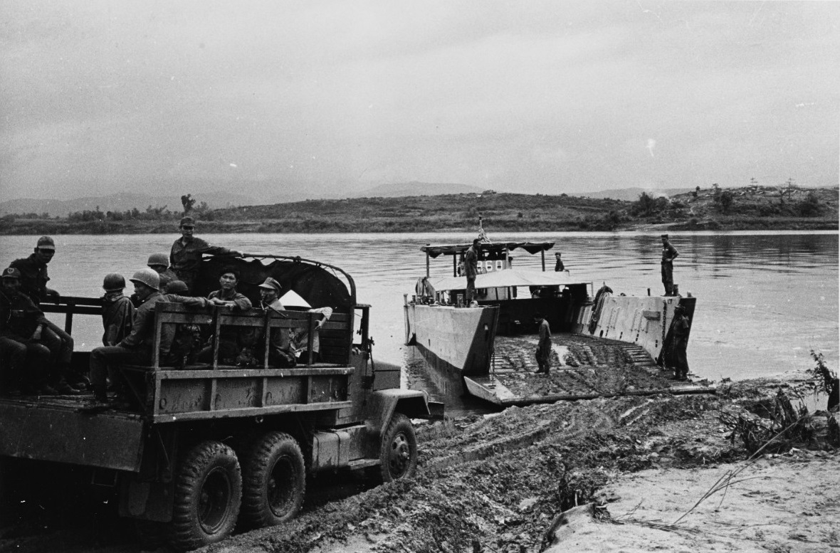 A landing craft numbered 860 nosed onto a sand bank, its ramp down, a truck loaded with soldiers waiting to drive aboard.