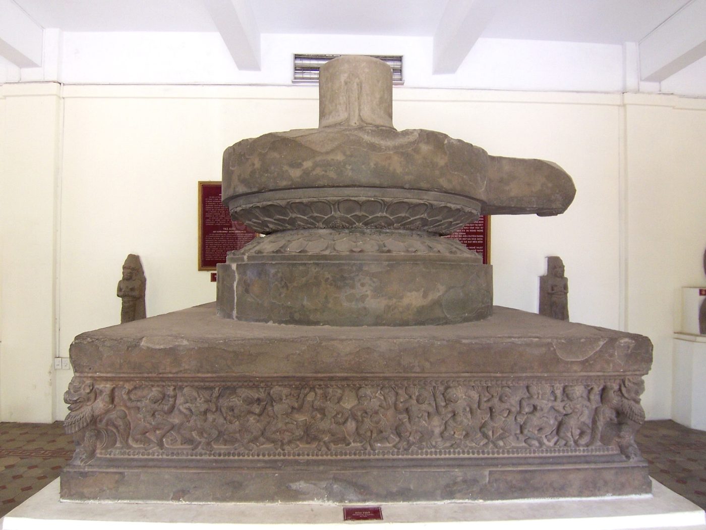 The Trà Kiệu pedestal, its stone face carved with a row of dancing apsaras and musicians, grey stone, shown in a museum.